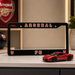 Black Arsenal FC license plate frame with a red car model on a shelf.