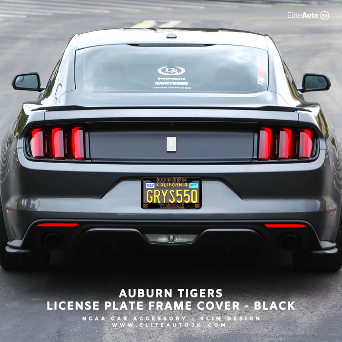 Rear view of a dark gray Mustang with a custom black license plate frame cover installed, featuring an Auburn Tigers themed California license plate. Text overlay at the bottom reads 'AUBURN TIGERS LICENSE PLATE FRAME COVER - BLACK' with 'EliteAuto' branding.
