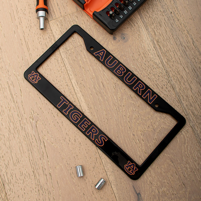 A black license plate frame with Auburn Tigers branding, laid flat on a light wood surface with mounting hardware nearby.