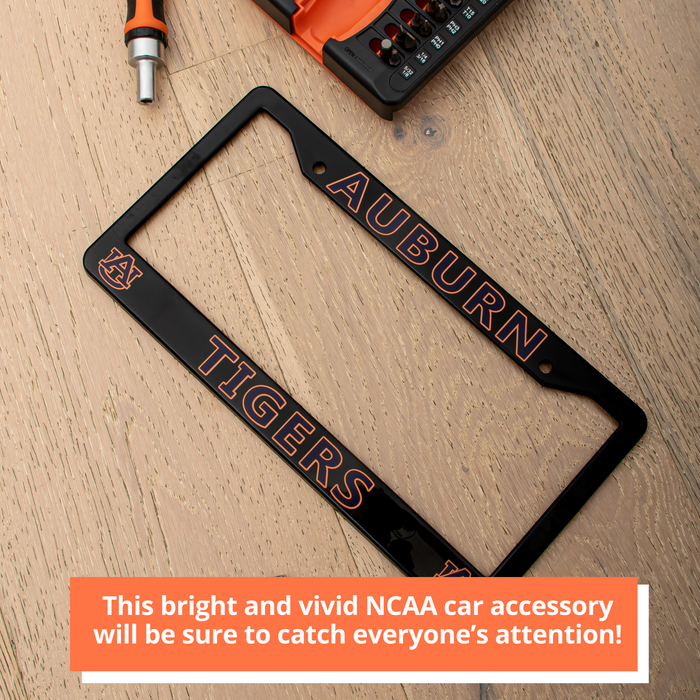 A black license plate frame with orange and white Auburn Tigers branding, displayed diagonally on a light wood background with a power drill partially visible at the top.