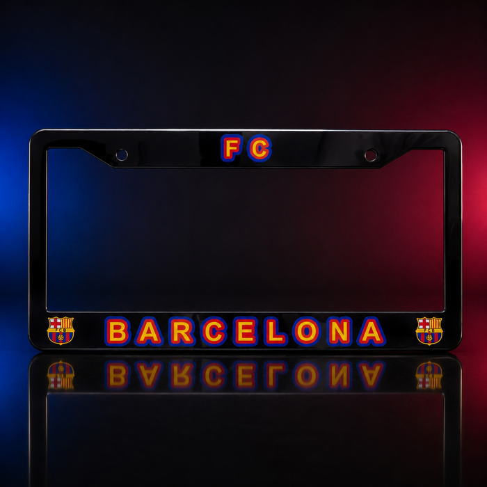 A black license plate frame featuring 'FC BARCELONA' in orange and blue lettering, with FC Barcelona club crests on the lower corners, shown against a dark studio background with red and blue lighting.