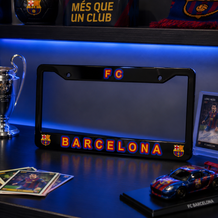 A black license plate frame featuring 'FC BARCELONA' in red and blue lettering, displayed on a dark shelf with blue LED lighting alongside soccer memorabilia.
