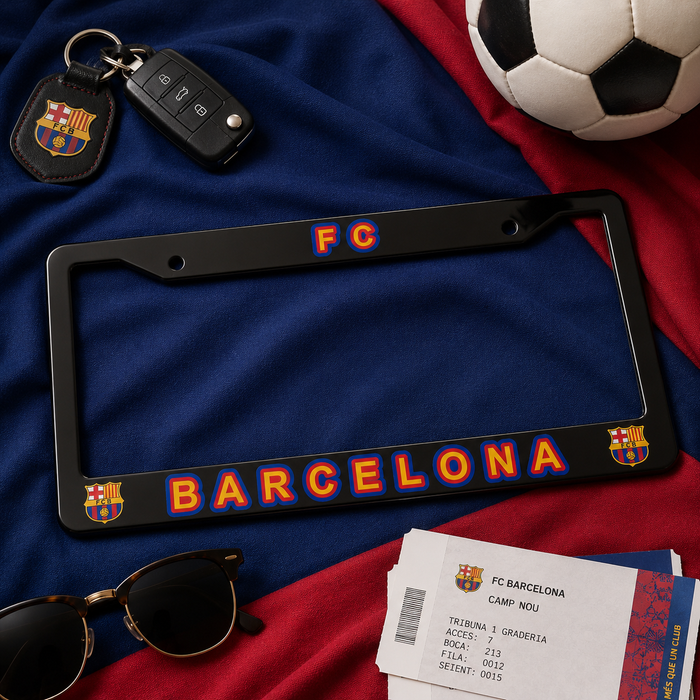 A black license plate frame with FC Barcelona branding, featuring 'FC' in orange and blue at the top and 'BARCELONA' in orange and blue along the bottom, flanked by club crests. The frame is styled in a flat-lay arrangement on a blue and red fabric background with a soccer ball, sunglasses, a car key fob, and a ticket.