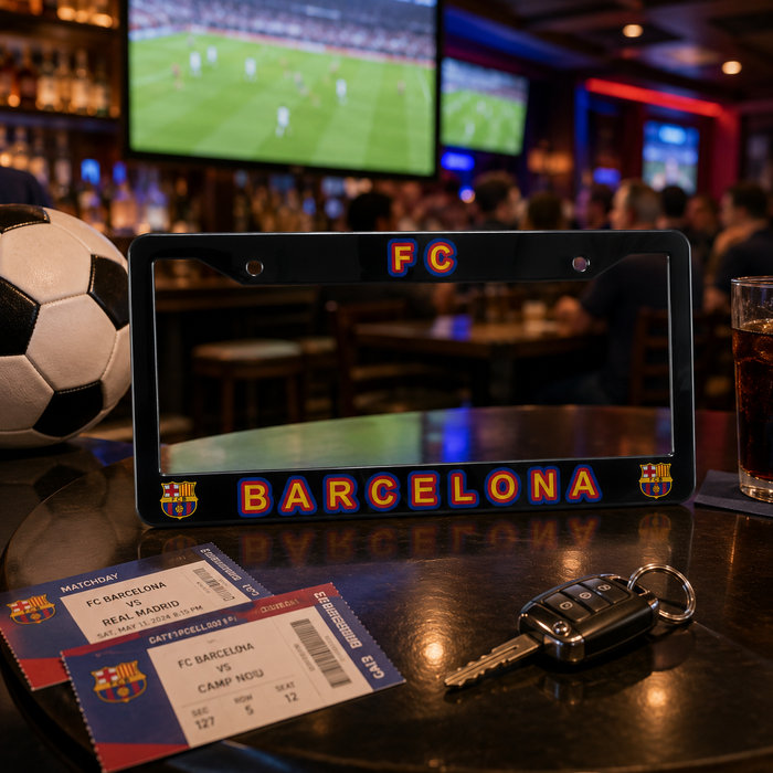 A black license plate frame featuring 'FC BARCELONA' in orange and blue lettering, displayed on a dark bar counter alongside a soccer ball, tickets, keys, and a drink, with a blurred bar interior and football match screens in the background.