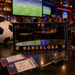 A black license plate frame featuring 'FC BARCELONA' in orange and blue lettering, displayed on a dark bar counter alongside a soccer ball, tickets, keys, and a drink, with a blurred bar interior and football match screens in the background.