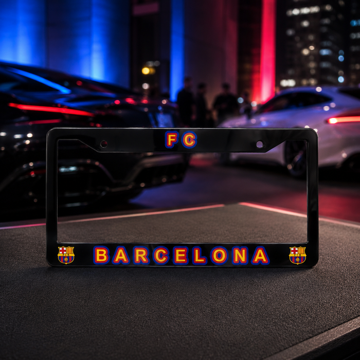 A black license plate frame featuring 'FC BARCELONA' in glowing orange and blue lettering, with the FC Barcelona crest on each corner, displayed on a dark surface with a blurred city street background.