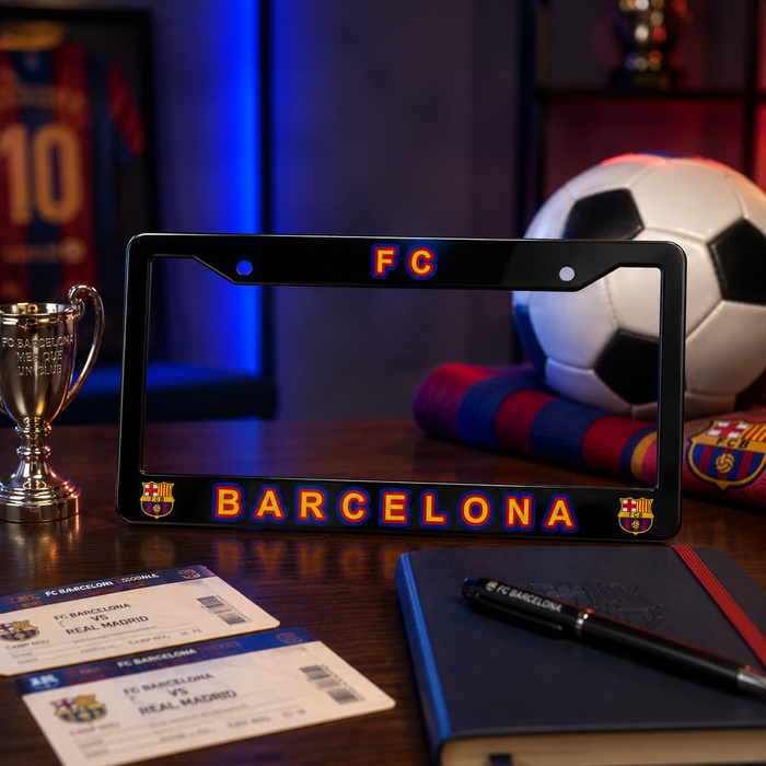 A black license plate frame featuring 'FC BARCELONA' in orange and blue lettering with club crests on the corners, displayed on a wooden table with soccer-themed props including a scarf, soccer ball, tickets, notebook, and pen.