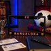 A black license plate frame featuring 'FC BARCELONA' in orange and blue lettering with club crests on the corners, displayed on a wooden table with soccer-themed props including a scarf, soccer ball, tickets, notebook, and pen.