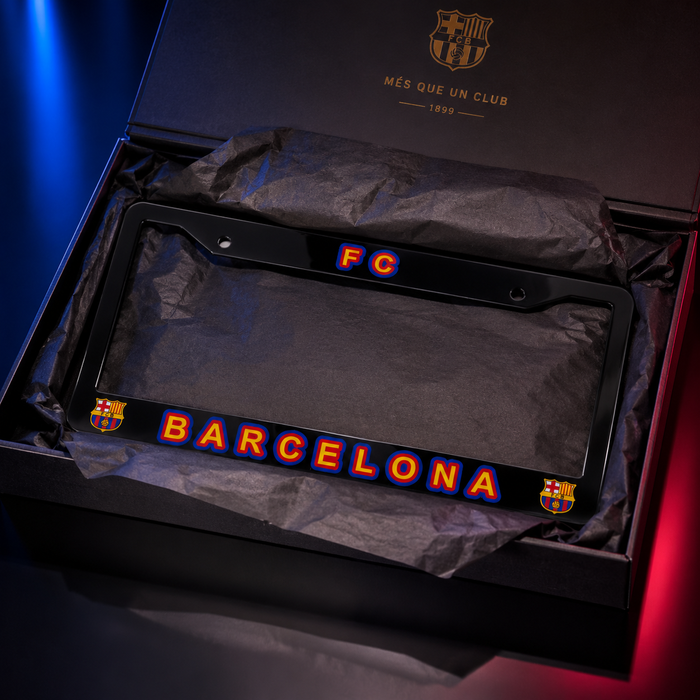 A black FC Barcelona license plate frame with 'FC' and 'BARCELONA' in red and blue lettering, displayed inside a branded black gift box with tissue paper. The FC Barcelona crest is visible on the frame and inside the box lid.