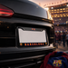 A black license plate frame with 'FC BARCELONA' in red and yellow lettering, mounted on the rear of a black car at a stadium during dusk. An FC Barcelona scarf is draped in the lower right corner.