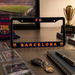 A black license plate frame featuring 'FC BARCELONA' in orange and blue lettering, displayed on a wooden desk surrounded by Barcelona-themed memorabilia including a scarf, framed soccer photos, a key, a notebook, and a trophy.