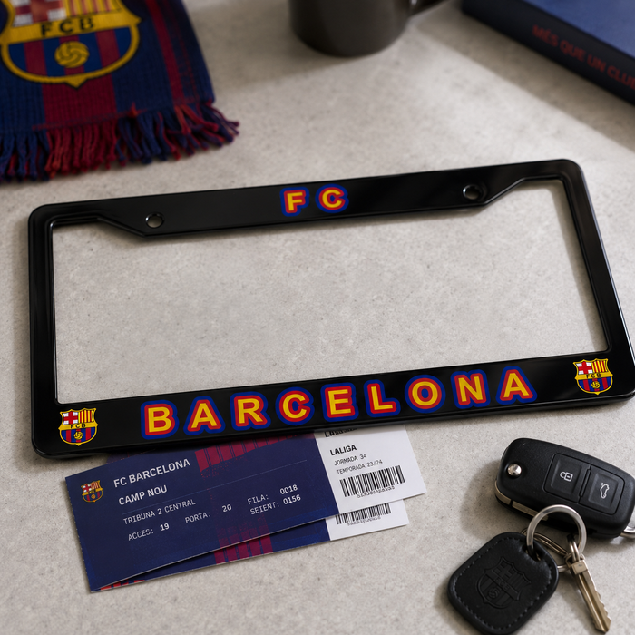 A black license plate frame featuring the FC Barcelona logo and 'BARCELONA' text in red, yellow, and blue, displayed on a light gray surface with an FC Barcelona ticket, keys, and a scarf nearby.
