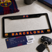 A black license plate frame featuring the FC Barcelona logo and 'BARCELONA' text in red, yellow, and blue, displayed on a light gray surface with an FC Barcelona ticket, keys, and a scarf nearby.