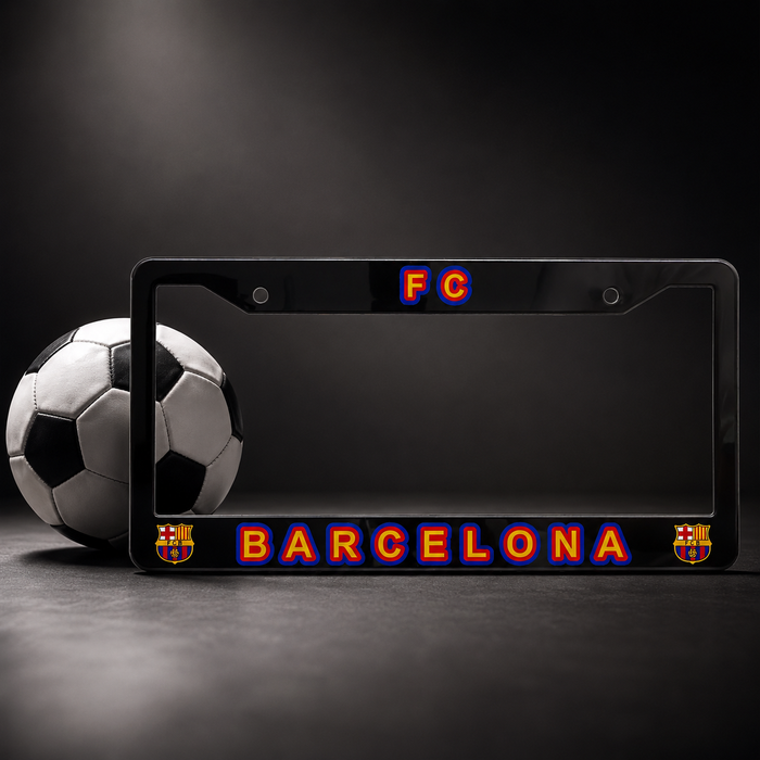 A black license plate frame featuring 'FC BARCELONA' in red and blue outlined lettering, with FC Barcelona crests on the corners, shown next to a soccer ball on a dark studio background.