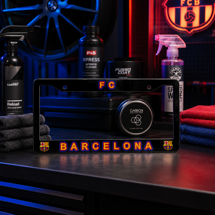A black license plate frame featuring the FC Barcelona logo and 'BARCELONA' text in glowing orange-red lettering, displayed on a dark workbench with car care products and towels in the background.