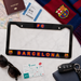 A black license plate frame with colorful FC Barcelona lettering and crest, styled in a flat lay with a passport, ticket, scarf, sunglasses, card, and key fob on a white marble surface.