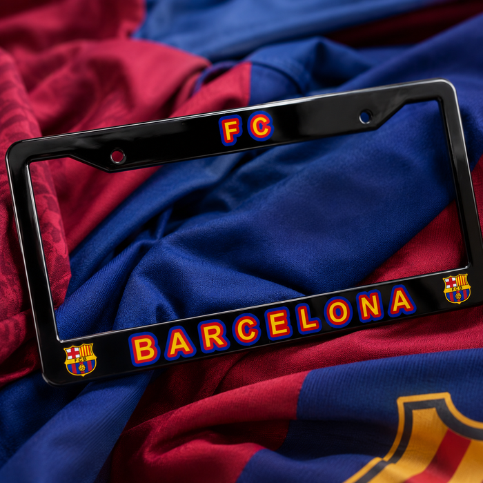 A black license plate frame featuring 'FC BARCELONA' in red and yellow with blue outlines, resting on a draped FC Barcelona jersey.