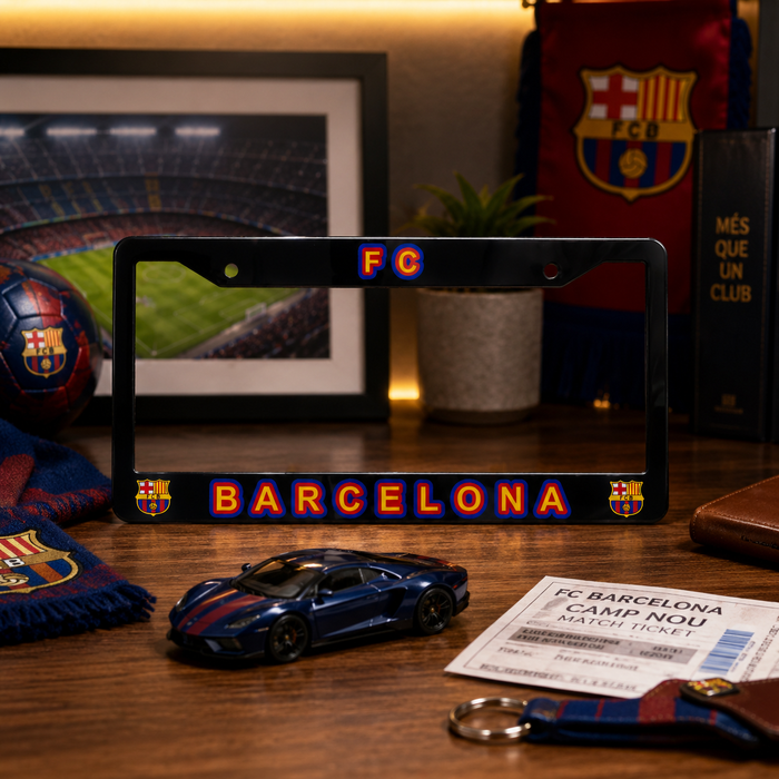 A black license plate frame featuring 'FC BARCELONA' in red and blue gradient letters, displayed on a wooden table with FC Barcelona-themed items including a scarf, toy car, ticket, and framed stadium photo.
