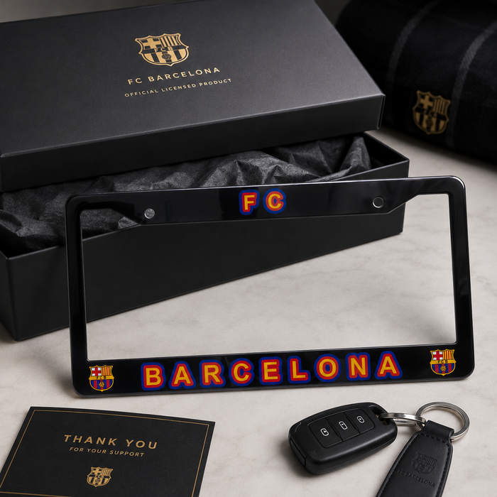 A black FC Barcelona license plate frame with colorful 'FC' and 'BARCELONA' lettering, displayed with a key fob, a thank you card, and the product box on a light marble surface.