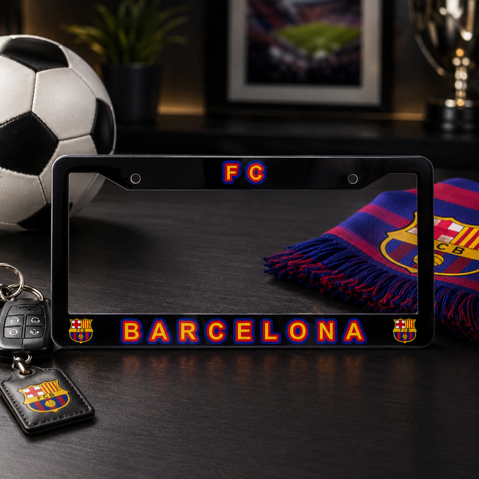 A black license plate frame featuring FC Barcelona branding, including 'FC' at the top and 'BARCELONA' with team crests at the bottom, displayed on a dark tabletop with a soccer ball, keys, and a scarf nearby.