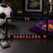 A black license plate frame featuring FC Barcelona branding, including 'FC' at the top and 'BARCELONA' with team crests at the bottom, displayed on a dark tabletop with a soccer ball, keys, and a scarf nearby.