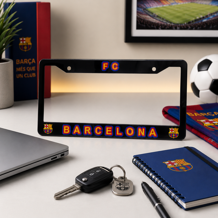 A black license plate frame featuring the FC Barcelona logo and 'FC BARCELONA' text in red, orange, and blue, displayed on a desk with sports-themed items including a notebook, keys, a laptop, and a soccer ball.