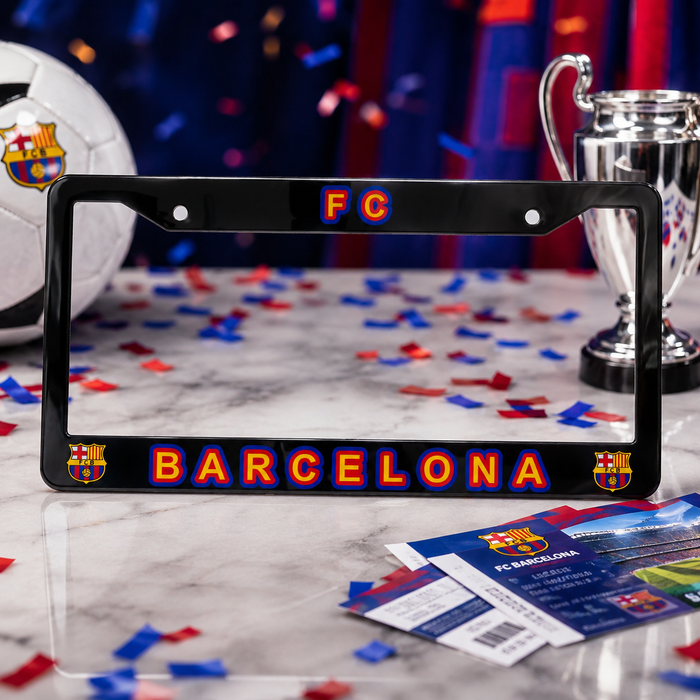 A black license plate frame featuring the FC Barcelona logo and 'BARCELONA' text in red and blue, displayed on a marble surface with soccer-themed props including a ball, trophy, and tickets.