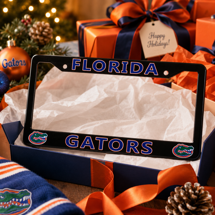 A black Florida Gators license plate frame displayed in a gift box with tissue paper, surrounded by holiday decorations including wrapped presents, an ornament, pinecones, and a scarf. The frame features 'FLORIDA' on the top and 'GATORS' on the bottom in blue with white outlines, plus the Gators logo on each corner.