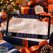 A black Florida Gators license plate frame displayed in a gift box with tissue paper, surrounded by holiday decorations including wrapped presents, an ornament, pinecones, and a scarf. The frame features 'FLORIDA' on the top and 'GATORS' on the bottom in blue with white outlines, plus the Gators logo on each corner.