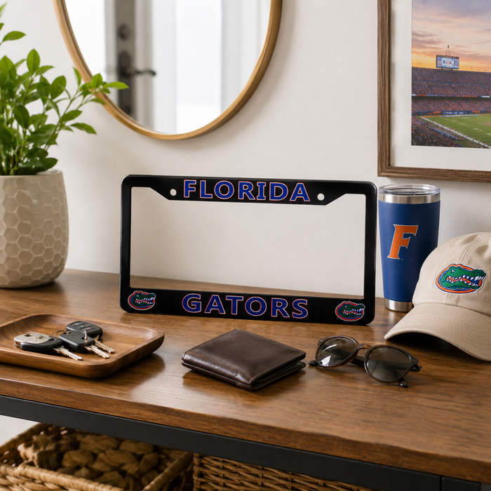 A black license plate frame with 'FLORIDA' and 'GATORS' in blue and orange Florida Gators lettering, styled on a wooden console table with a wallet, sunglasses, keys, a tumbler, and a cap.