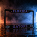 A black Florida Gators license plate frame with blue and purple lettering and alligator logos, shown on a wet reflective surface with dramatic orange and blue lighting and smoke in the background.