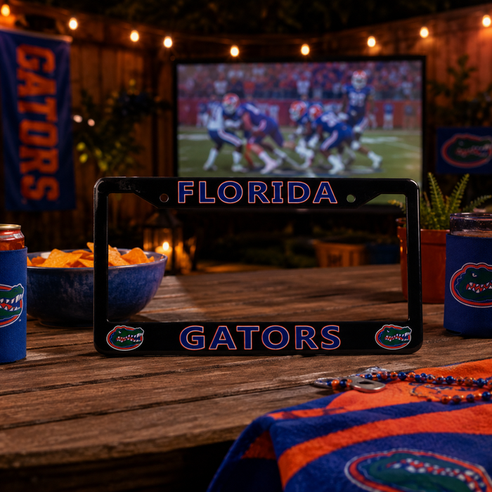 A black Florida Gators license plate frame displayed on a wooden table at a night game, surrounded by Florida Gators themed party decorations including a blue can, a bowl of chips, a TV showing a football play, and a blue and orange towel.