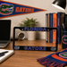 A black Florida Gators license plate frame displayed on a wooden desk, surrounded by themed Florida Gators decor including a banner, scarf, and books.