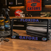 A black Florida Gators license plate frame displayed on a wooden workbench in a garage-like setting. The frame features 'FLORIDA' at the top and 'GATORS' at the bottom in blue letters with a white outline, plus Florida Gators logos in the lower corners.