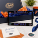 A Florida Gators themed license plate frame displayed on a white marble surface, surrounded by a navy blue gift box with gold foil, orange tissue paper, a 'Go Gators!' card, an orange envelope, a blue ribbon, and a white medallion.