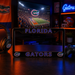 A black Florida Gators license plate frame with blue and orange lettering, displayed on a dark wooden table in a themed room with gaming accessories and a TV showing a football game.