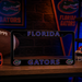 A black Florida Gators license plate frame with blue and purple lettering, displayed on a wooden table with Florida Gators themed decor in the background.