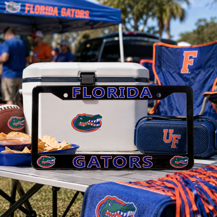 A black Florida Gators license plate frame displayed on a folding table at a game, surrounded by Florida Gators merchandise including a cooler, chair, towel, and snacks.