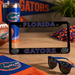 A black Florida Gators license plate frame displayed on a wooden table, surrounded by Florida-themed merchandise including a tumbler, football, sunglasses, and a scarf.