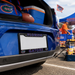 A close-up of a blue car trunk with Florida Gators branding, including a license plate frame that says 'FLORIDA GATORS' with team logos, a cooler, a football, and a bag in the background at an outdoor event.