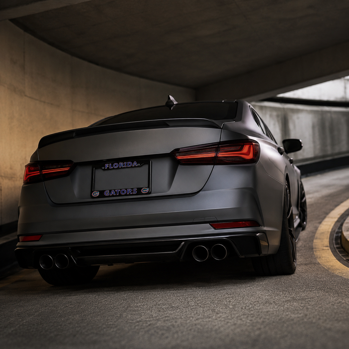 Rear three-quarter view of a matte dark gray sedan parked under a concrete tunnel, showing the back of the car with dual exhausts, a Florida/Gators license plate, and illuminated LED taillights.