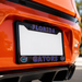 Close-up of a black Florida Gators license plate frame mounted on the rear bumper of an orange car. The frame features 'FLORIDA' at the top and 'GATORS' at the bottom in blue outlined letters, with Florida Gators logos on the lower corners.