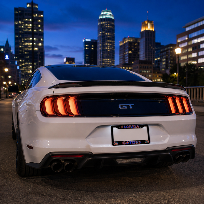 Rear view of a white Ford Mustang GT with illuminated LED taillights and a black rear spoiler, parked outdoors at dusk with a city skyline in the background.