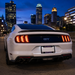 Rear view of a white Ford Mustang GT with illuminated LED taillights and a black rear spoiler, parked outdoors at dusk with a city skyline in the background.