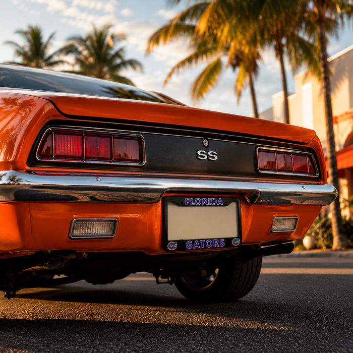 Close-up rear view of an orange classic car with 'SS' badging on the trunk, parked outdoors on asphalt with palm trees and buildings in the background. A Florida license plate with 'GATORS' is visible.