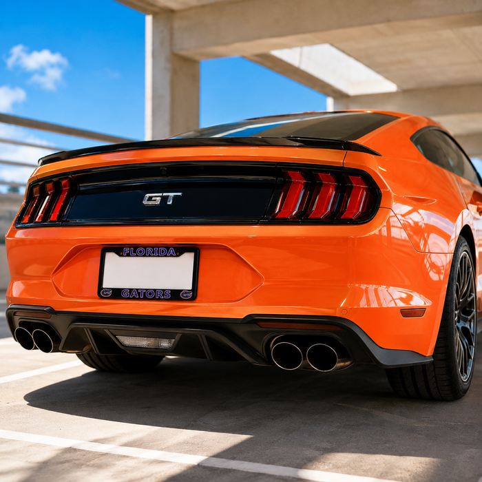 Rear three-quarter view of a bright orange Ford Mustang GT parked in a concrete parking structure under a blue sky. The car features a black rear spoiler, black GT badge, black rear window, black trim, and dual chrome exhaust tips.