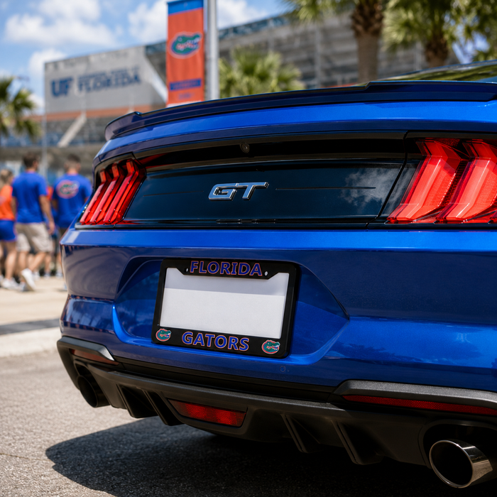 Close-up rear view of a blue Ford Mustang GT with a Florida Gators license plate frame and Florida license plate, parked outdoors near a stadium with palm trees and people blurred in the background.
