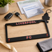 Black license plate frame with 'Liverpool FC' on a wooden surface with keys and a driver guide.