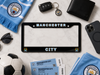 A black license plate frame with 'MANCHESTER CITY' in white and blue lettering is displayed in a flat-lay arrangement with Manchester City themed accessories, including a scarf, tickets, a soccer ball, sunglasses, a wallet, a phone case, and a key fob, on a white marble background.