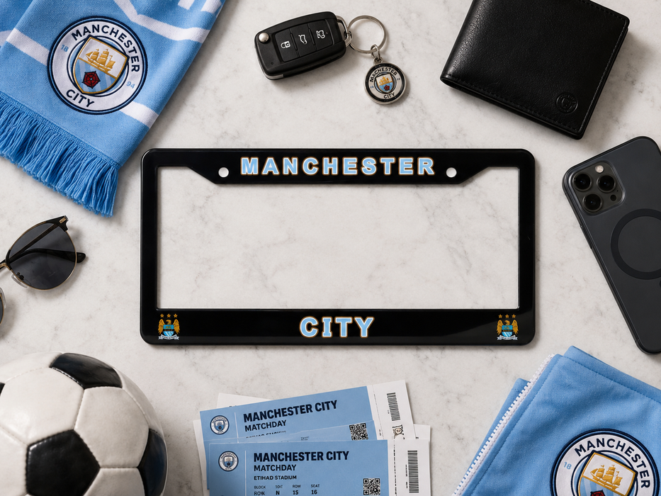 A black license plate frame with 'MANCHESTER CITY' in white and blue lettering is displayed in a flat-lay arrangement with Manchester City themed accessories, including a scarf, tickets, a soccer ball, sunglasses, a wallet, a phone case, and a key fob, on a white marble background.